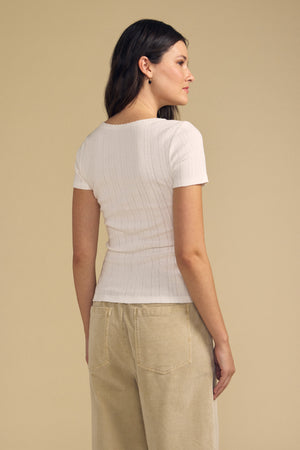 A woman stands sideways, facing right, wearing the LENA POINTELLE TEE by Velvet by Graham & Spencer—a white ribbed tee with a fitted silhouette—paired with beige pants against a plain beige background.