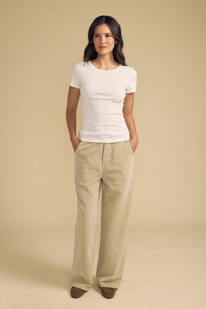 A woman stands with her hands in her pockets, wearing the LENA POINTELLE TEE by Velvet by Graham & Spencer and loose beige pants, set against a plain beige background.