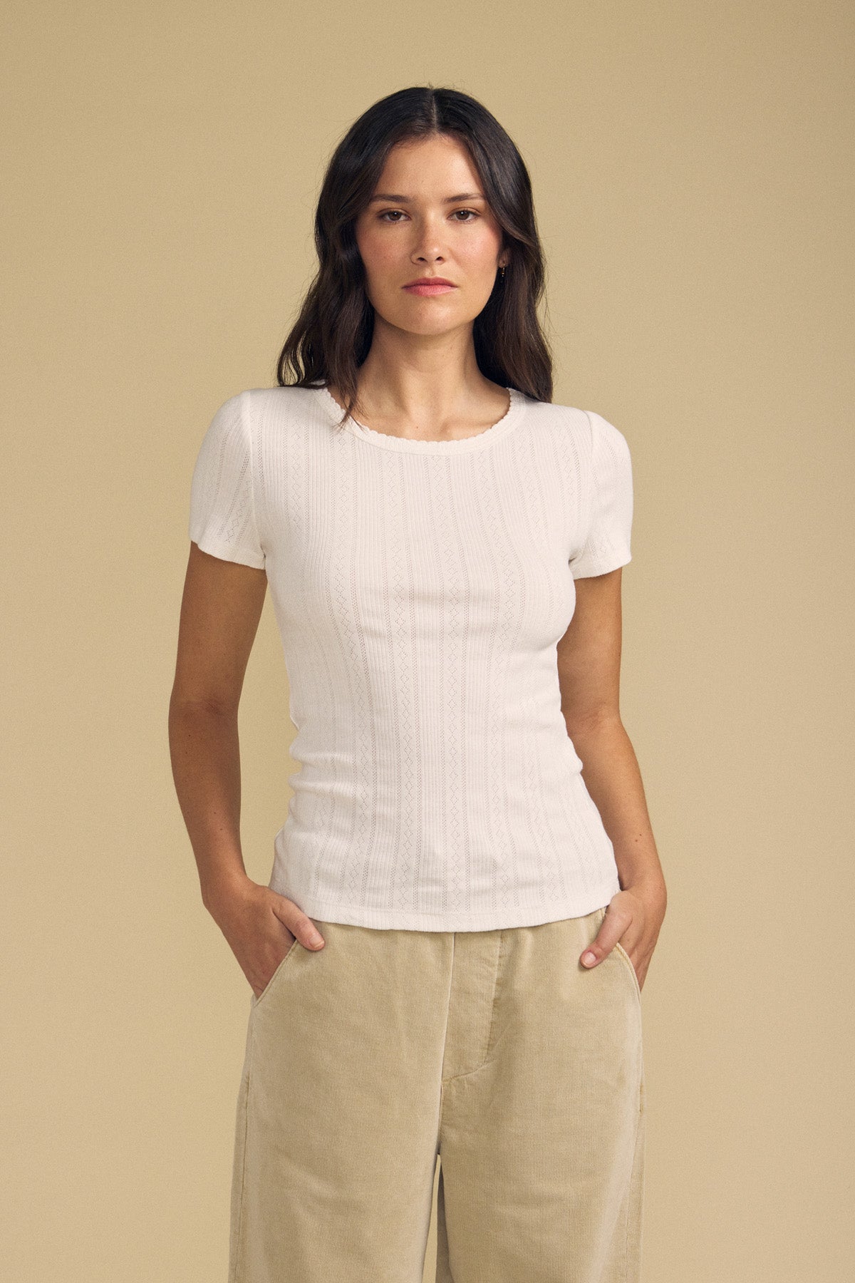 A woman stands against a beige background with her hands in her pockets and a neutral expression, wearing the LENA POINTELLE TEE by Velvet by Graham & Spencer—a fitted silhouette cotton knit short-sleeve white top—paired with tan pants.-41834091610305