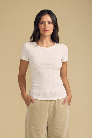 A woman stands against a beige background with her hands in her pockets and a neutral expression, wearing the LENA POINTELLE TEE by Velvet by Graham & Spencer—a fitted silhouette cotton knit short-sleeve white top—paired with tan pants.