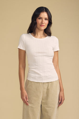 A woman wearing the LENA POINTELLE TEE by Velvet by Graham & Spencer in white, paired with beige pants, stands against a plain beige background.