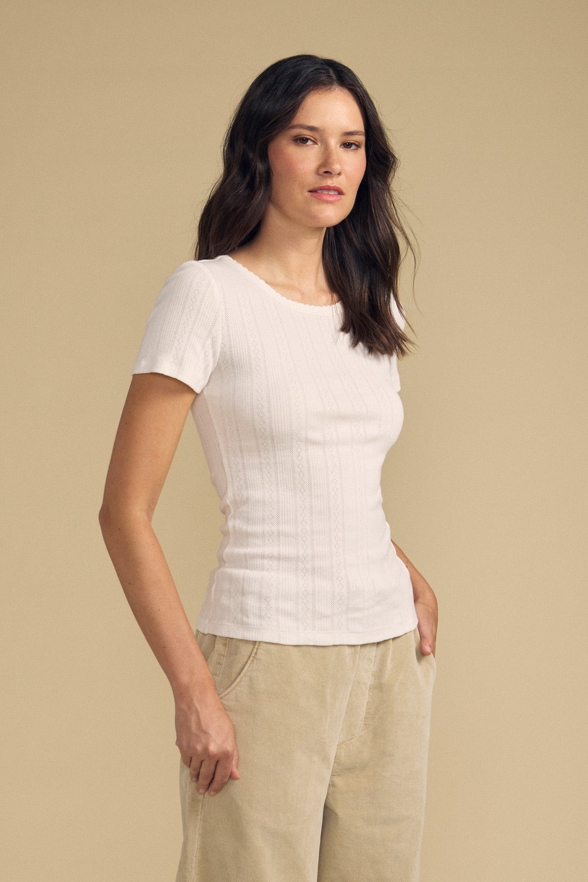 A woman stands against a beige background wearing the fitted silhouette LENA POINTELLE TEE by Velvet by Graham & Spencer—a short-sleeve white cotton knit ribbed top—paired with light beige pants, one hand in her pocket.