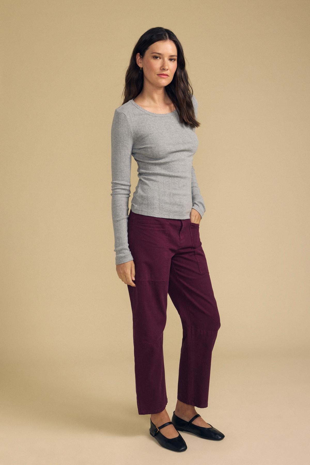 A person stands against a beige background wearing the MIRIAM TEE by Velvet by Graham & Spencer—a gray cotton knit long-sleeve shirt featuring a scalloped crew neckline—paired with burgundy pants and black shoes, one hand casually tucked in their pocket.