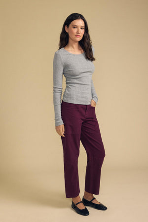 A person stands against a beige background wearing the MIRIAM TEE by Velvet by Graham & Spencer—a gray cotton knit long-sleeve shirt featuring a scalloped crew neckline—paired with burgundy pants and black shoes, one hand casually tucked in their pocket.