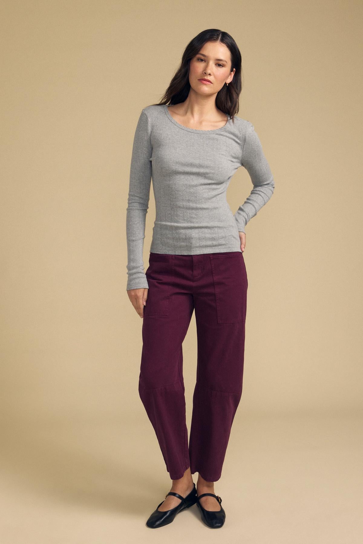 A woman stands against a beige background, wearing a long-sleeve gray top and the BRYLIE PANT by Velvet by Graham & Spencer—structured cotton twill barrel leg trousers in burgundy with utilitarian patch pockets—paired with black flats, one hand behind her back.-42091995431105