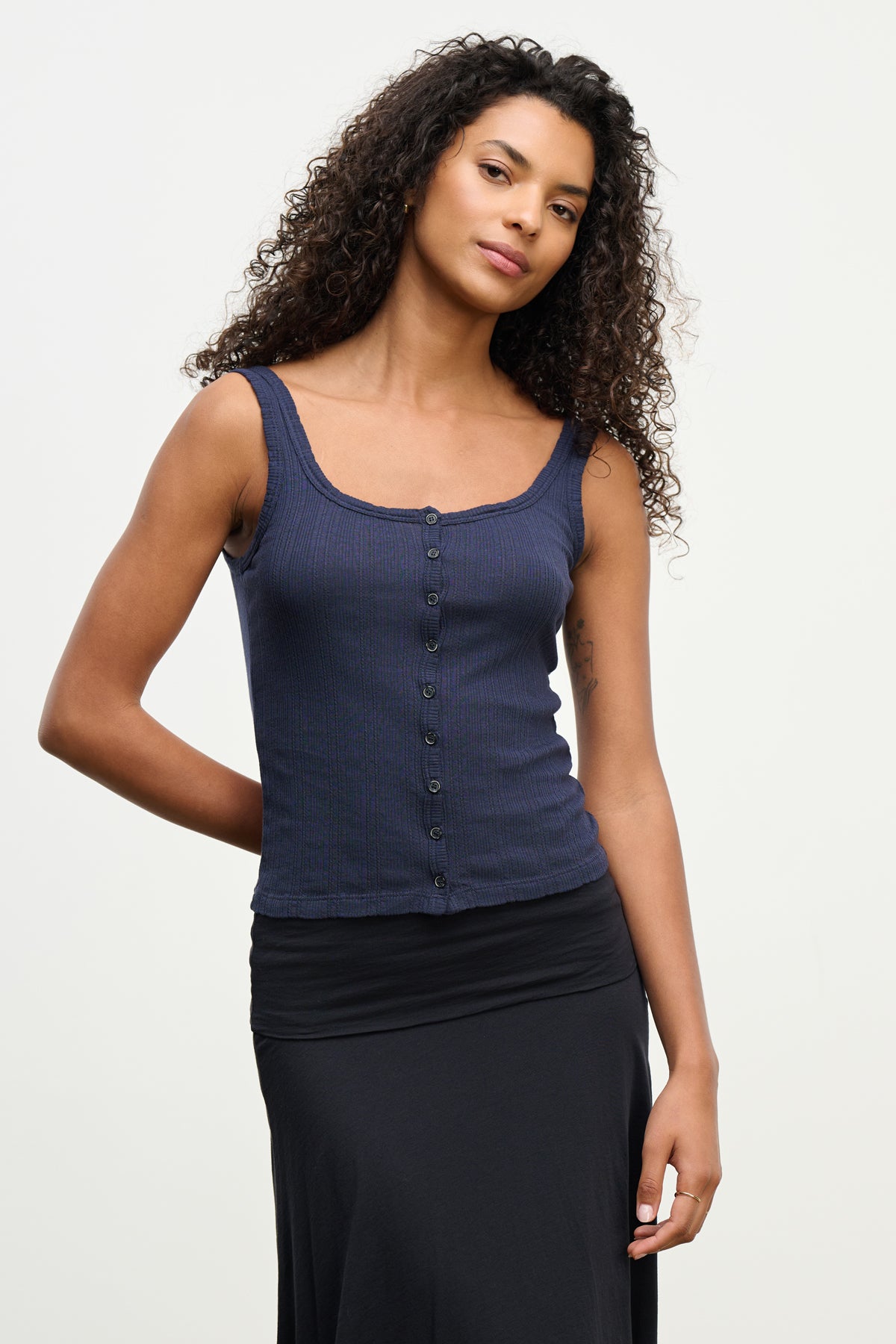 A woman with curly hair models the ADELLE TANK TOP by Velvet by Graham & Spencer in navy, paired with a black skirt, standing before a plain light background.-39538872615105
