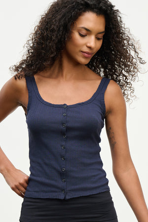 A woman with curly hair wears the ADELLE TANK TOP by Velvet by Graham & Spencer, a sleeveless, dark blue fitted style, paired with black bottoms against a plain light background.