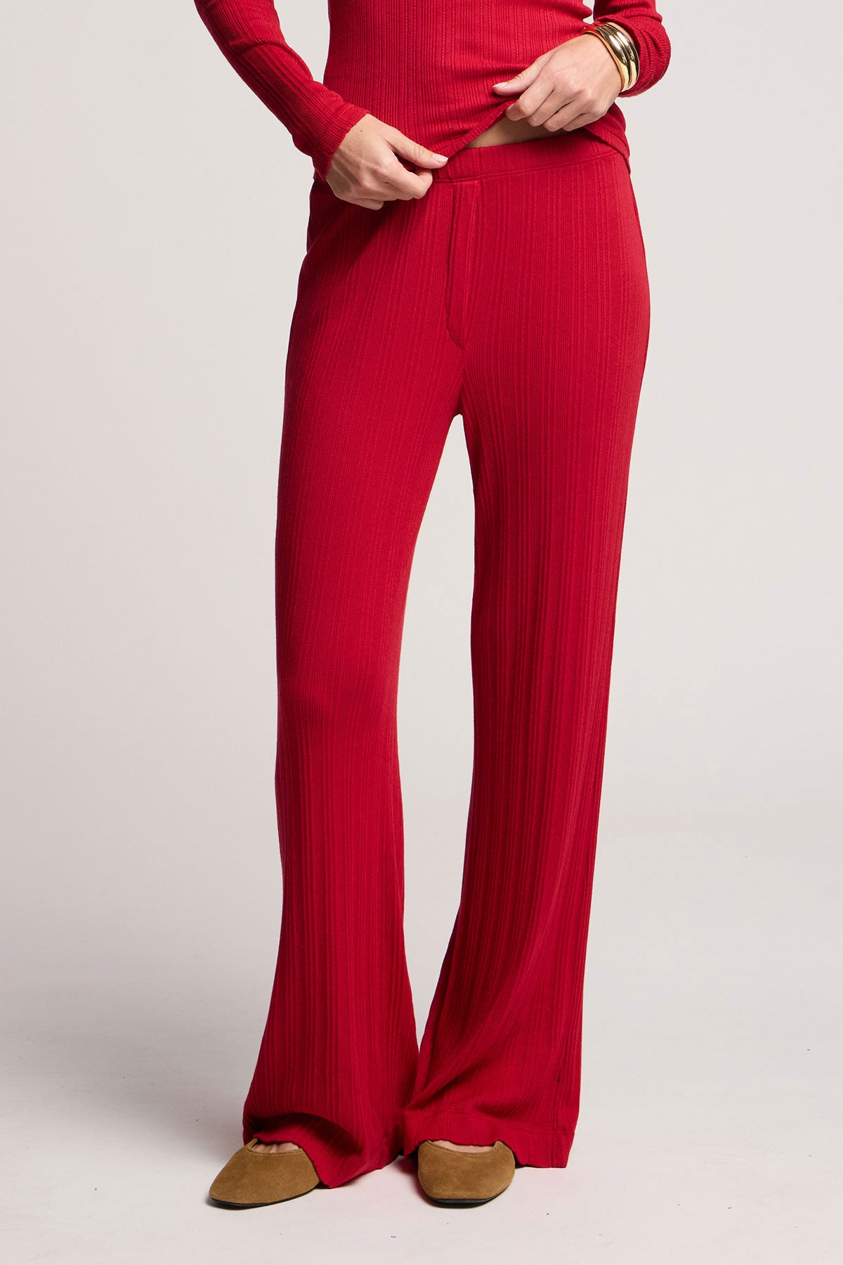 Wearing Velvet by Graham & Spencer’s ALEMI POINTELLE PANT in red with ribbed pointelle accents and a matching long-sleeve top, gold bracelets, and tan slippers, the person stands against a plain light background.