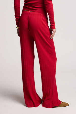 Person wearing Velvet by Graham & Spencer's ALEMI POINTELLE PANT, a high-waisted, flared leg red pant, paired with a matching long-sleeve top, stands facing away from the camera with one arm at their side.