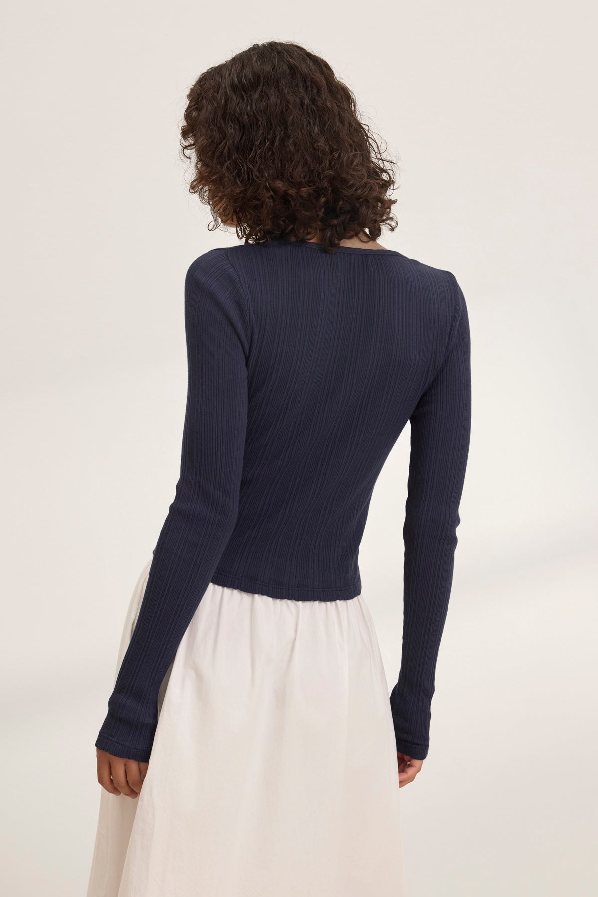 The individual elegantly showcases layering by wearing Velvet by Graham & Spencer's navy DREW POINTELLE CARDIGAN and a white skirt, standing with their back to the camera against a plain backdrop.-38802080727233
