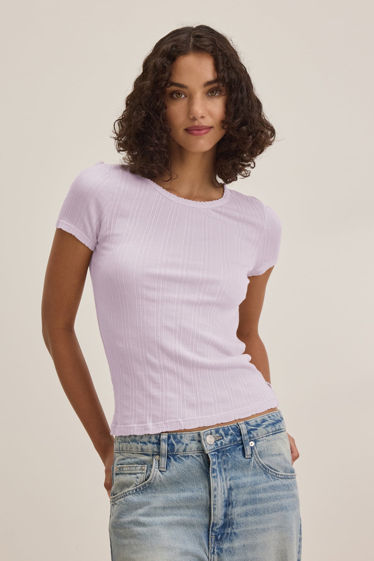 A woman wears the ENATI POINTELLE TEE by Velvet by Graham & Spencer, a light purple short-sleeve ribbed top with scalloped edges, paired with light blue jeans, standing against an off-white background.-39173111709889