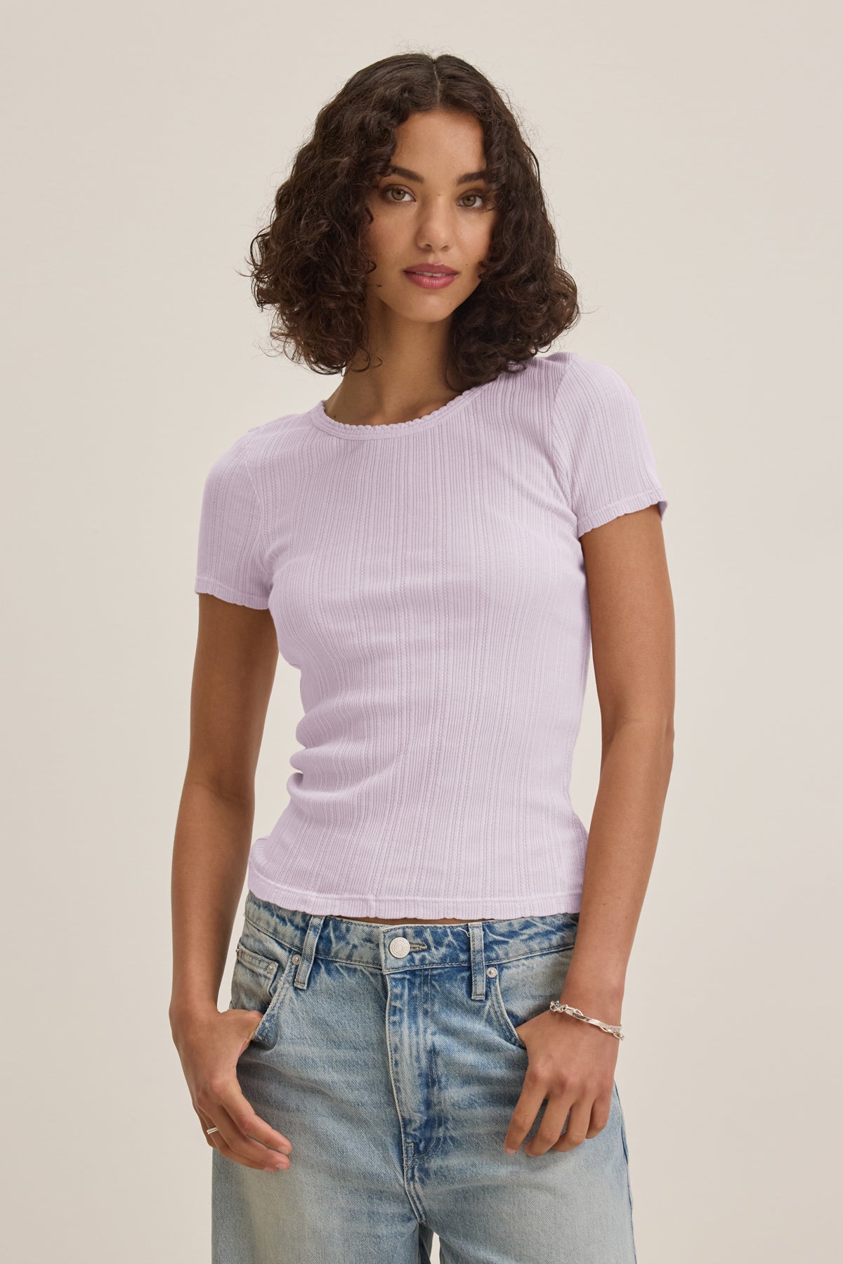 A woman with curly brown hair wears the Velvet by Graham & Spencer ENATI POINTELLE TEE in light purple, featuring ribbed pointelle details, paired with light blue jeans, standing against a plain off-white background.-39173111742657