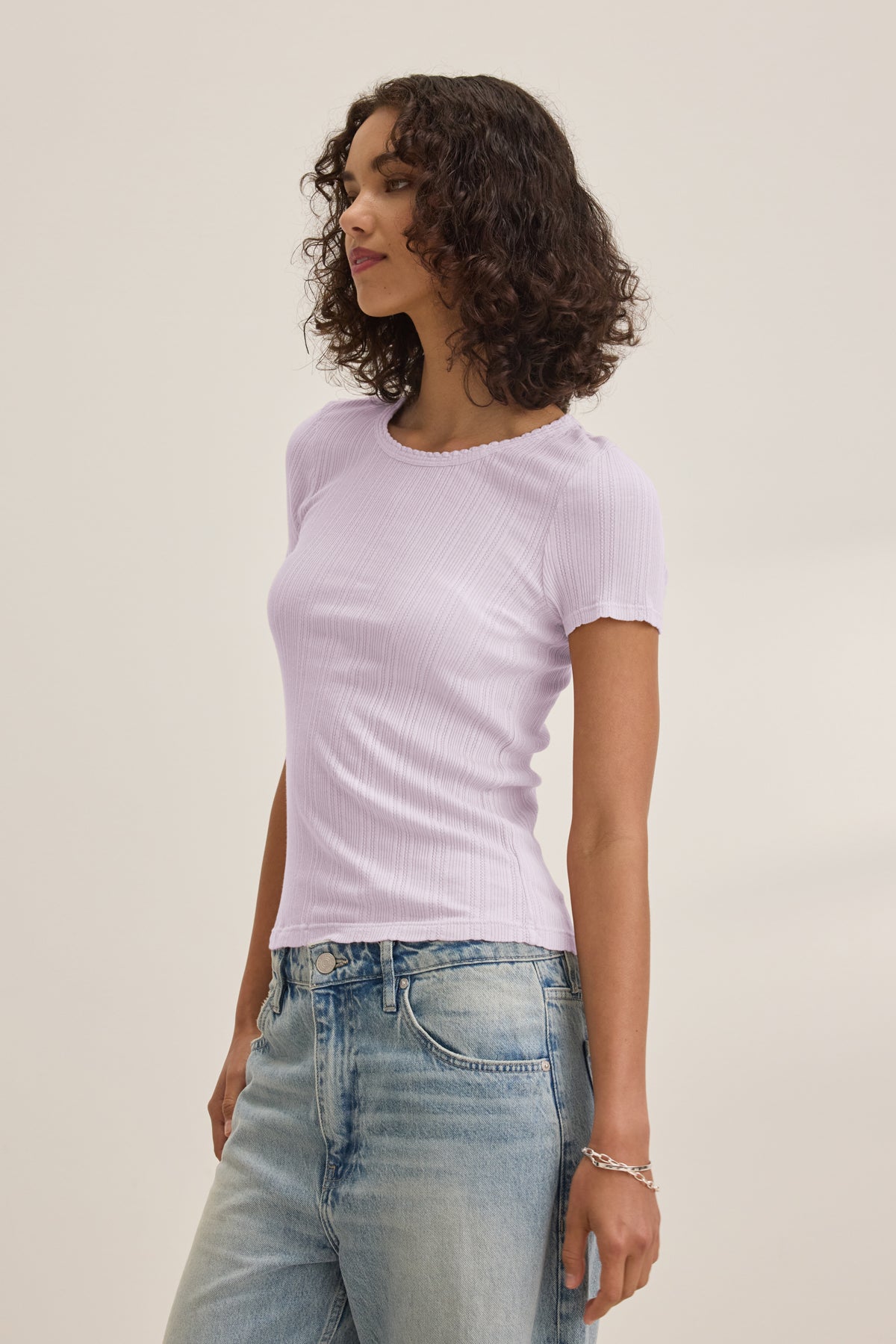   A woman wears the ENATI POINTELLE TEE by Velvet by Graham & Spencer—a light purple ribbed fitted top in a soft cotton-modal blend—paired with light blue jeans, standing against a plain light background. 
