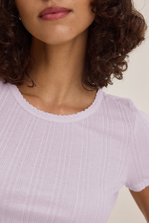 A woman with curly brown hair is wearing the ENATI POINTELLE TEE by Velvet by Graham & Spencer, a light purple fitted ribbed cotton-modal top with a scalloped neckline. Only her lower face, neck, and upper torso are visible.
