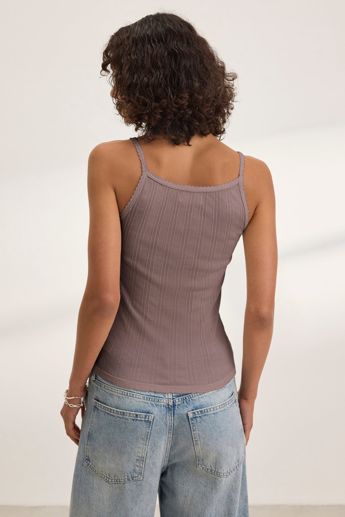 A person with curly hair faces away, wearing the Velvet by Graham & Spencer INIKU POINTELLE TANK TOP in mauve and light blue jeans in a minimal indoor setting.-39173124128961