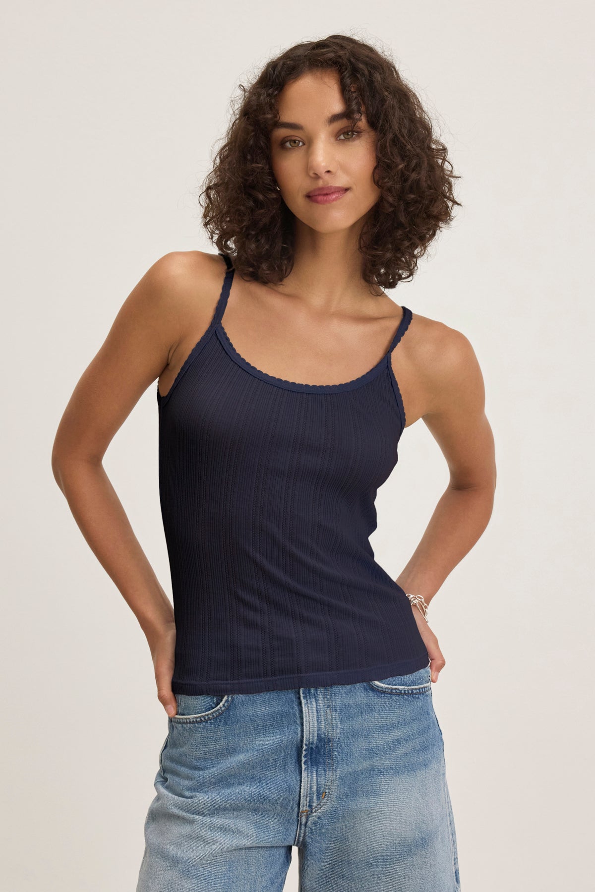   A woman with curly hair wears the Velvet by Graham & Spencer INIKU POINTELLE TANK TOP and light blue jeans, standing against a plain light background with her hands in her pockets. 