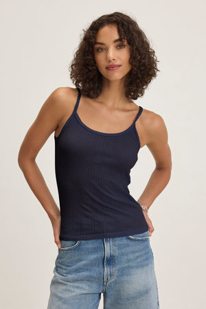 A woman with curly hair wears the Velvet by Graham & Spencer INIKU POINTELLE TANK TOP and light blue jeans, standing against a plain light background with her hands in her pockets.