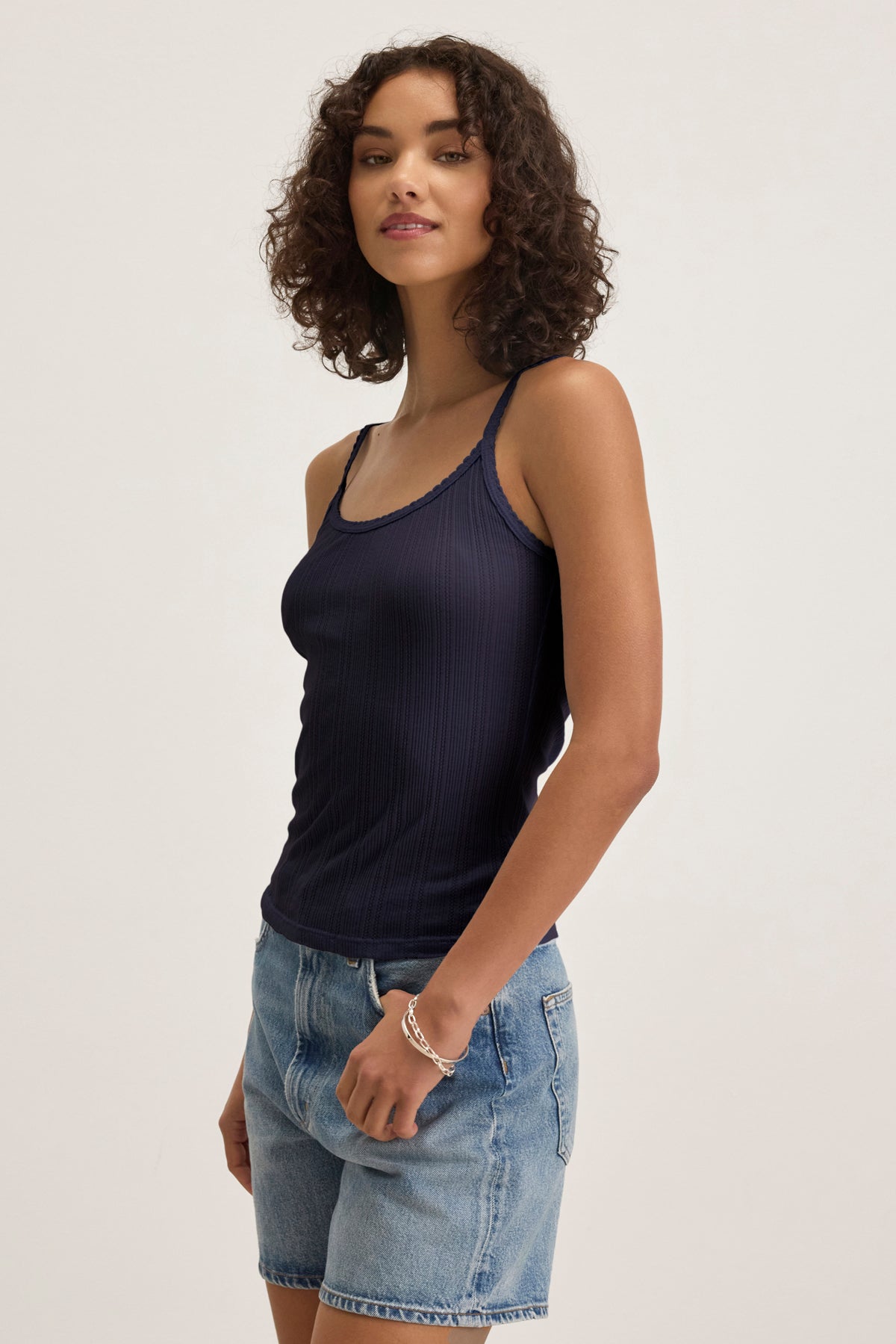 A woman with short curly hair wears the INIKU POINTELLE TANK TOP by Velvet by Graham & Spencer, a navy blue cotton-modal blend tank with scalloped edges, paired with denim shorts against a plain light background.-39173124325569