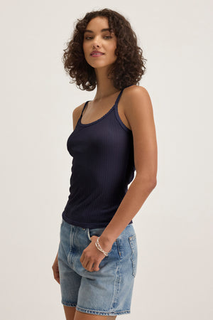 A woman with short curly hair wears the INIKU POINTELLE TANK TOP by Velvet by Graham & Spencer, a navy blue cotton-modal blend tank with scalloped edges, paired with denim shorts against a plain light background.