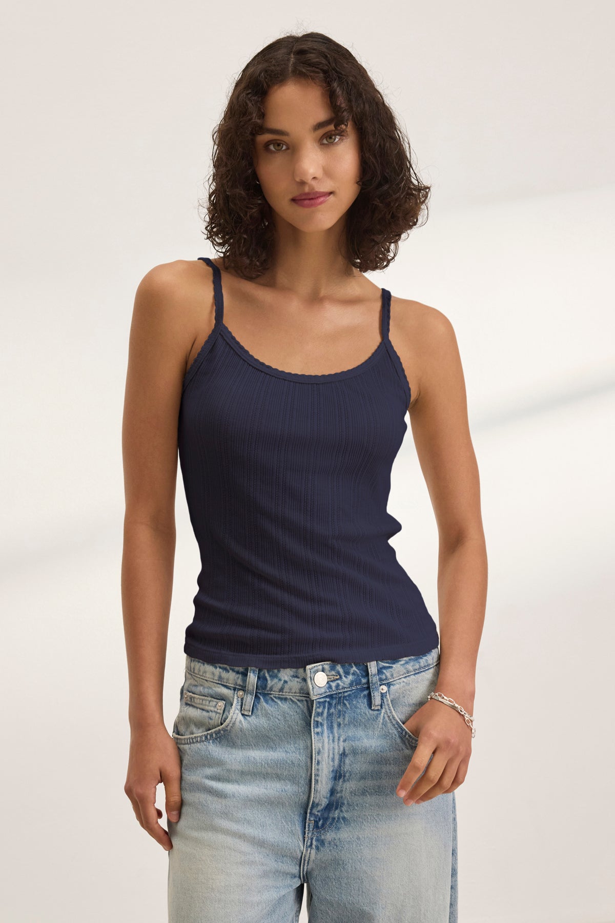 A woman with curly dark hair wears the INIKU POINTELLE TANK TOP by Velvet by Graham & Spencer—a navy cotton-modal blend tank with a ribbed texture—paired with light blue jeans, standing against a plain light background.-39173124358337