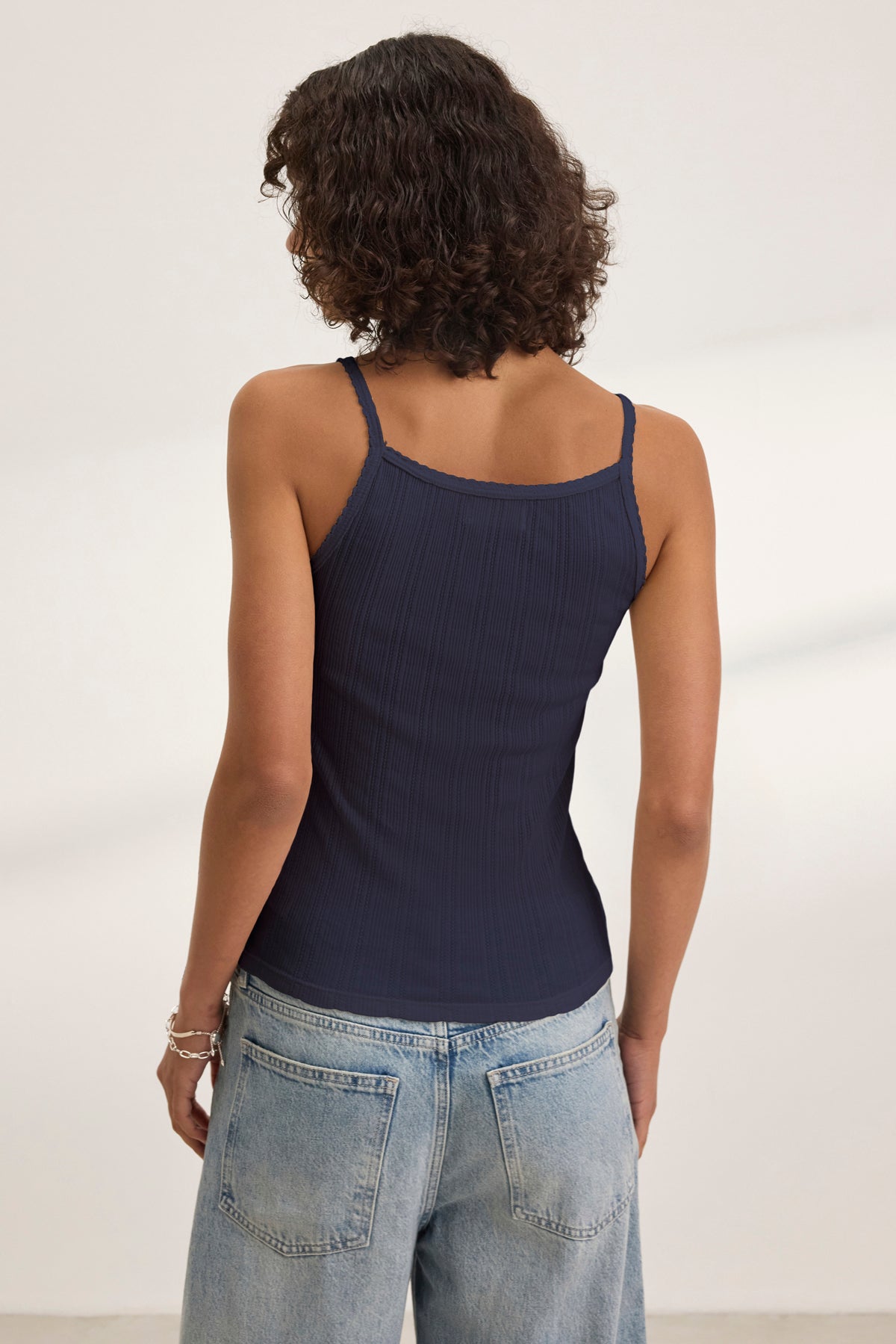 A person with curly brown hair, wearing the Velvet by Graham & Spencer INIKU POINTELLE TANK TOP in navy and light blue jeans, stands facing away from the camera in a minimal, light-colored room.-39173124391105