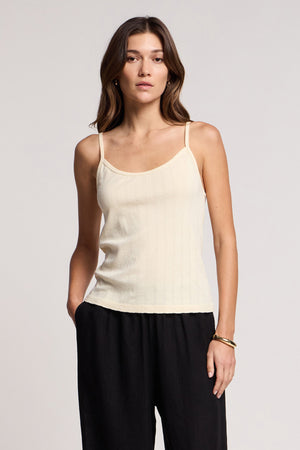 A woman stands against a neutral background, wearing the LYNN TANK TOP by Velvet by Graham & Spencer—a cream cotton tank with a fitted silhouette—paired with black pants and one hand in her pocket.