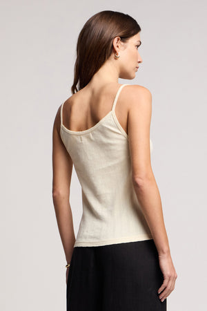 A woman faces away from the camera, wearing the LYNN TANK TOP by Velvet by Graham & Spencer—a cream cotton tank with a fitted silhouette—paired with black pants against a plain background.