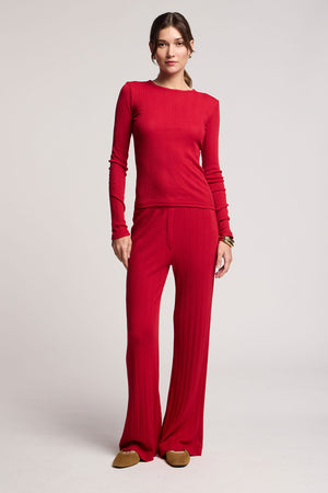 A woman stands facing forward in the ALEMI POINTELLE PANT and matching red long-sleeve top from Velvet by Graham & Spencer, styled with brown slippers and gold bangles, against a plain light background.
