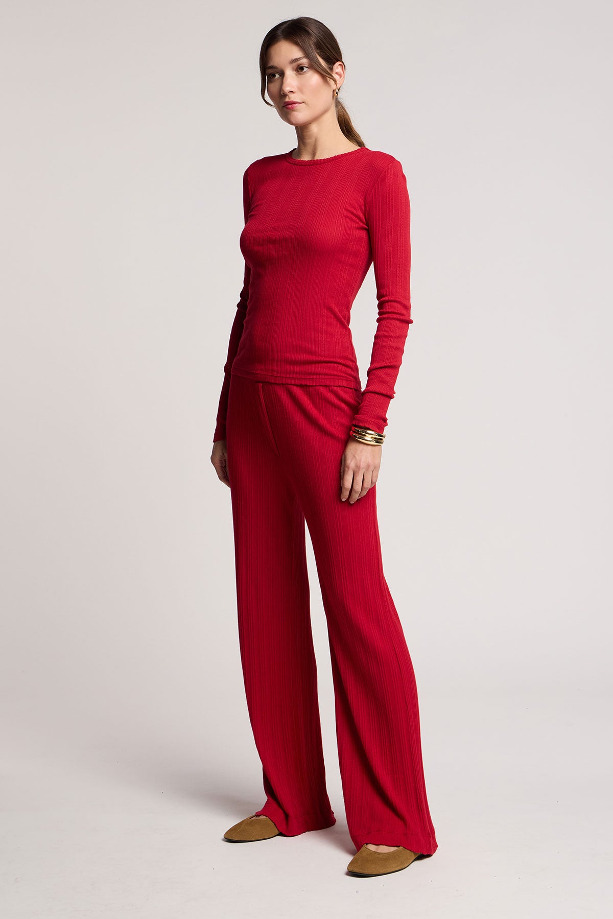 A woman stands against a plain light background wearing Velvet by Graham & Spencer’s ALEMI POINTELLE PANT, a matching long-sleeve red top, brown flat shoes, and gold bangles.