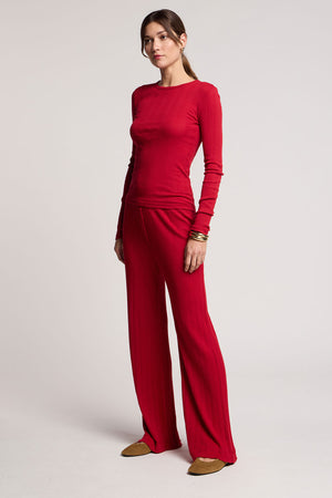 A woman stands against a plain light background wearing Velvet by Graham & Spencer’s ALEMI POINTELLE PANT, a matching long-sleeve red top, brown flat shoes, and gold bangles.