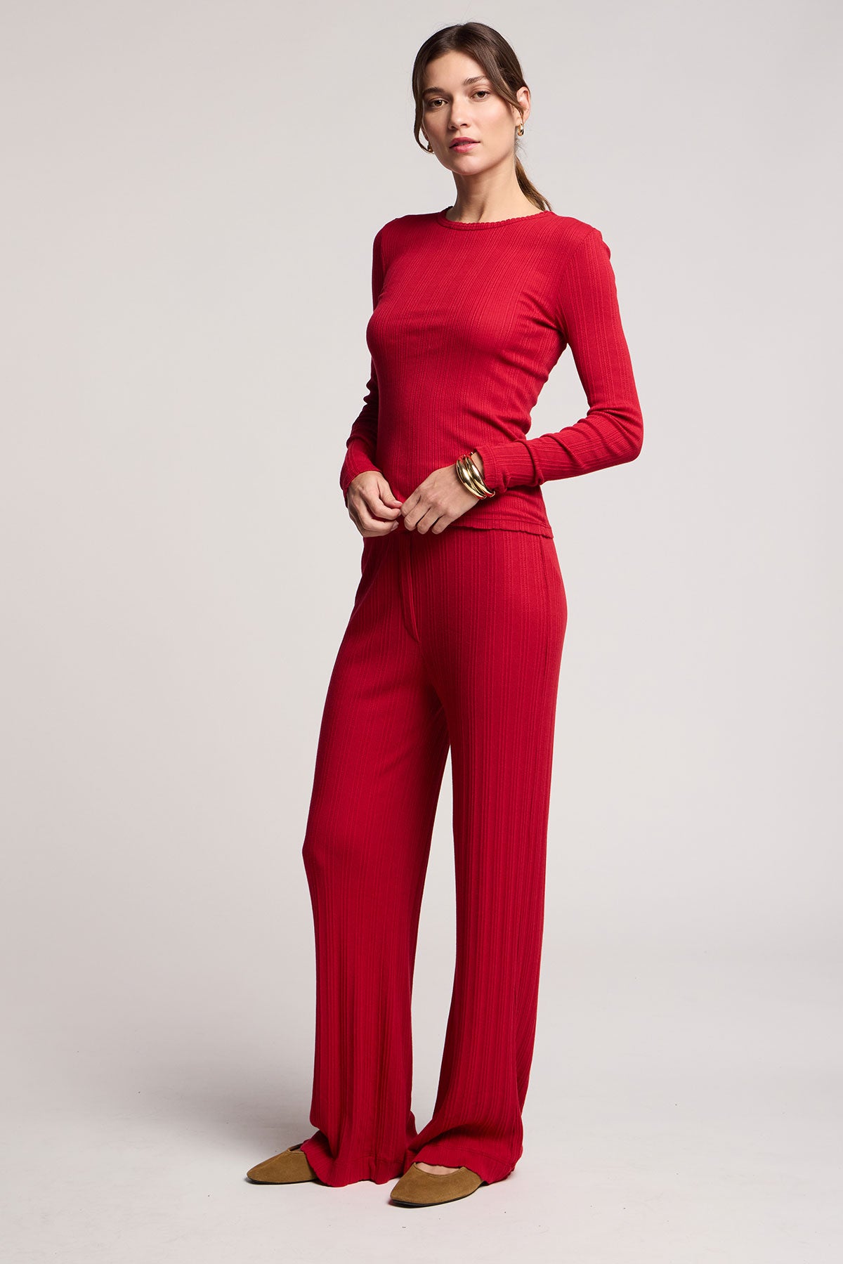 A woman poses against a plain light background, wearing the MAURI POINTELLE TEE by Velvet by Graham & Spencer—a matching red ribbed long-sleeve tee and wide-leg pant set—styled with bracelets and flat tan shoes.-40416831701185
