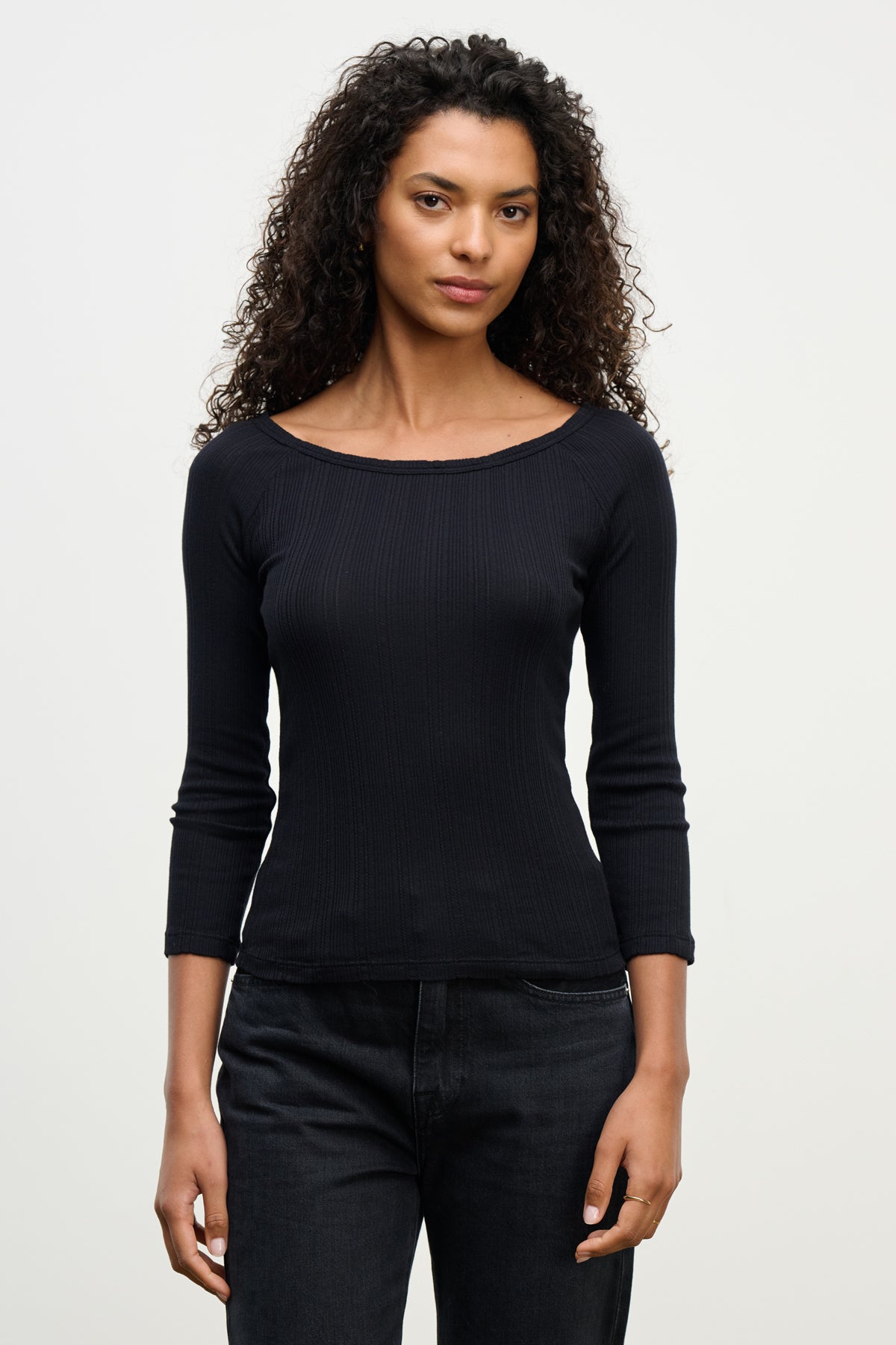 A woman with curly hair wears the Velvet by Graham & Spencer NIMA POINTELLE TEE, a fitted long-sleeve black boatneck top, with dark jeans, standing against a plain light background.-39742363238593