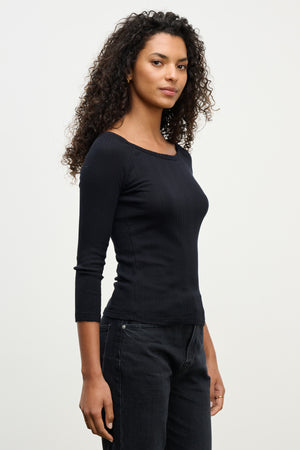 A woman with curly hair wears the Velvet by Graham & Spencer NIMA POINTELLE TEE in black and black jeans, standing against a plain light background, facing slightly to the side.