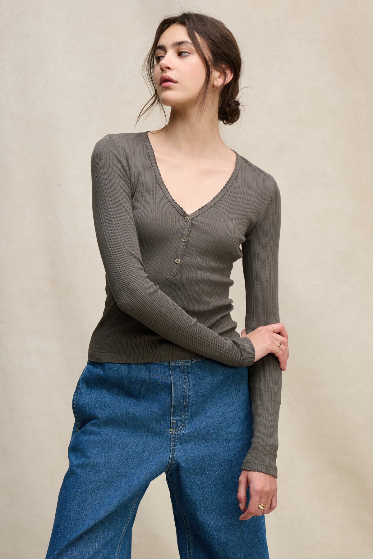 A person wears Velvet by Graham & Spencer's WINONA POINTELLE HENLEY—a ribbed, long-sleeve grey V-neck top—paired with loose blue jeans, standing against a beige background for an effortlessly feminine everyday look.-39742399348929
