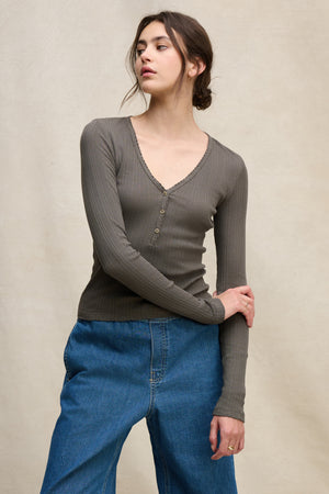 A person wears Velvet by Graham & Spencer's WINONA POINTELLE HENLEY—a ribbed, long-sleeve grey V-neck top—paired with loose blue jeans, standing against a beige background for an effortlessly feminine everyday look.