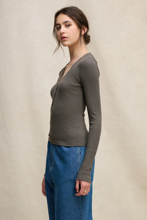 A woman with long brown hair in a low bun stands sideways against a neutral background, wearing the Velvet by Graham & Spencer WINONA POINTELLE HENLEY and blue jeans—a perfect feminine everyday layer.