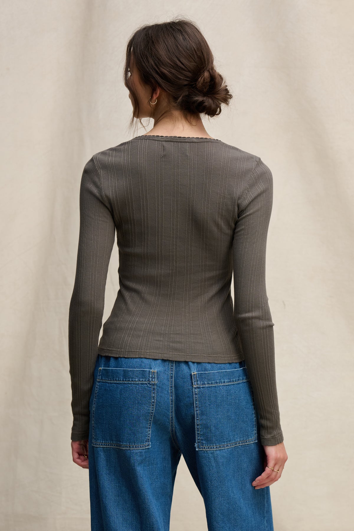 A person with brown hair in a loose bun is shown from the back wearing the WINONA POINTELLE HENLEY by Velvet by Graham & Spencer and blue jeans against a neutral background—a feminine everyday layer.-39742399283393