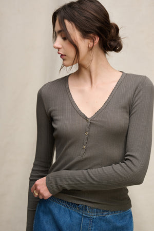 A woman with brown hair in a low bun wears the WINONA POINTELLE HENLEY by Velvet by Graham & Spencer, styled with blue jeans, standing against a neutral background.