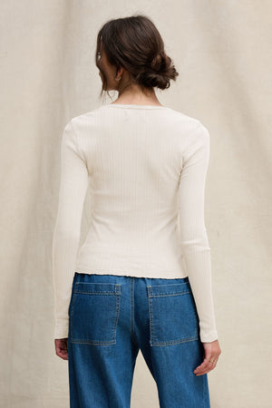 A woman stands facing away from the camera, wearing the WINONA POINTELLE HENLEY by Velvet by Graham & Spencer and blue denim jeans with large back pockets.