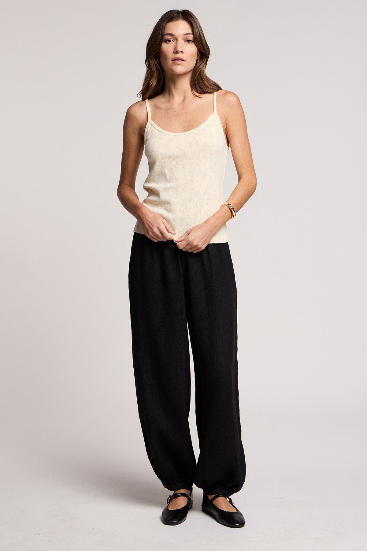 A woman stands against a plain background wearing Velvet by Graham & Spencer's CAMBER LINEN PANT in black with an elastic waist, paired with a beige sleeveless top and black flat shoes.-41371565228225