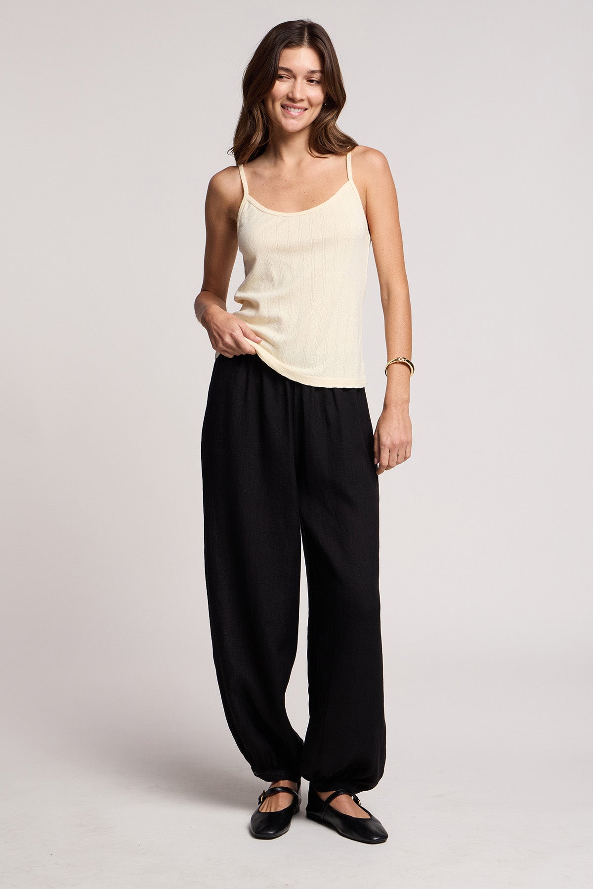 A woman stands smiling against a plain light background, wearing a sleeveless beige top, black CAMBER LINEN PANT by Velvet by Graham & Spencer with an elastic waist, black shoes, and a bracelet.