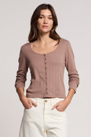 A woman faces forward, hands in her pockets, wearing the Velvet by Graham & Spencer ZELDA CARDIGAN—mauve cotton with ribbed texture and buttons—paired with white pants against a plain backdrop.