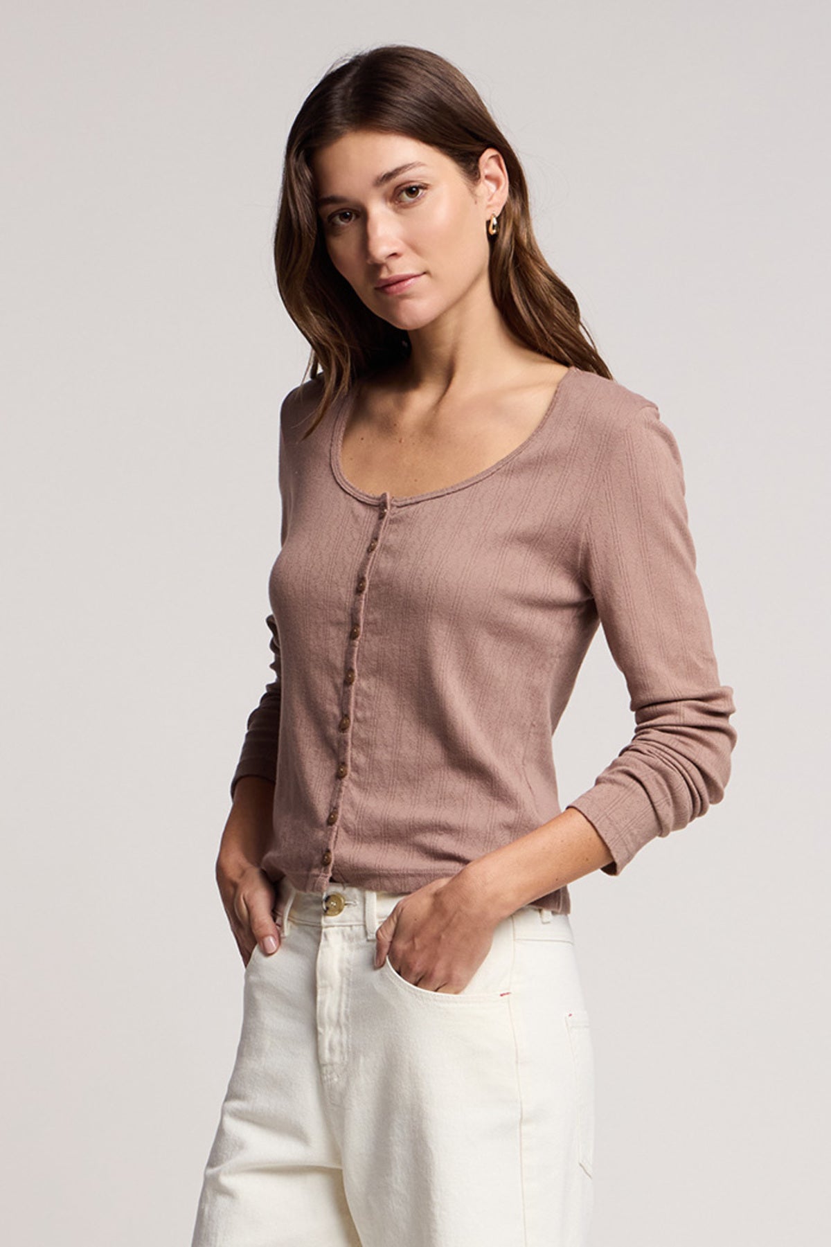 A woman with long brown hair models the ZELDA CARDIGAN by Velvet by Graham & Spencer—a mauve cotton piece with scallop cuffs—paired with white pants, set against a plain light background.