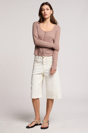 A woman stands against a plain background wearing the ZELDA CARDIGAN by Velvet by Graham & Spencer, styled with off-white knee-length culottes and black sandals.