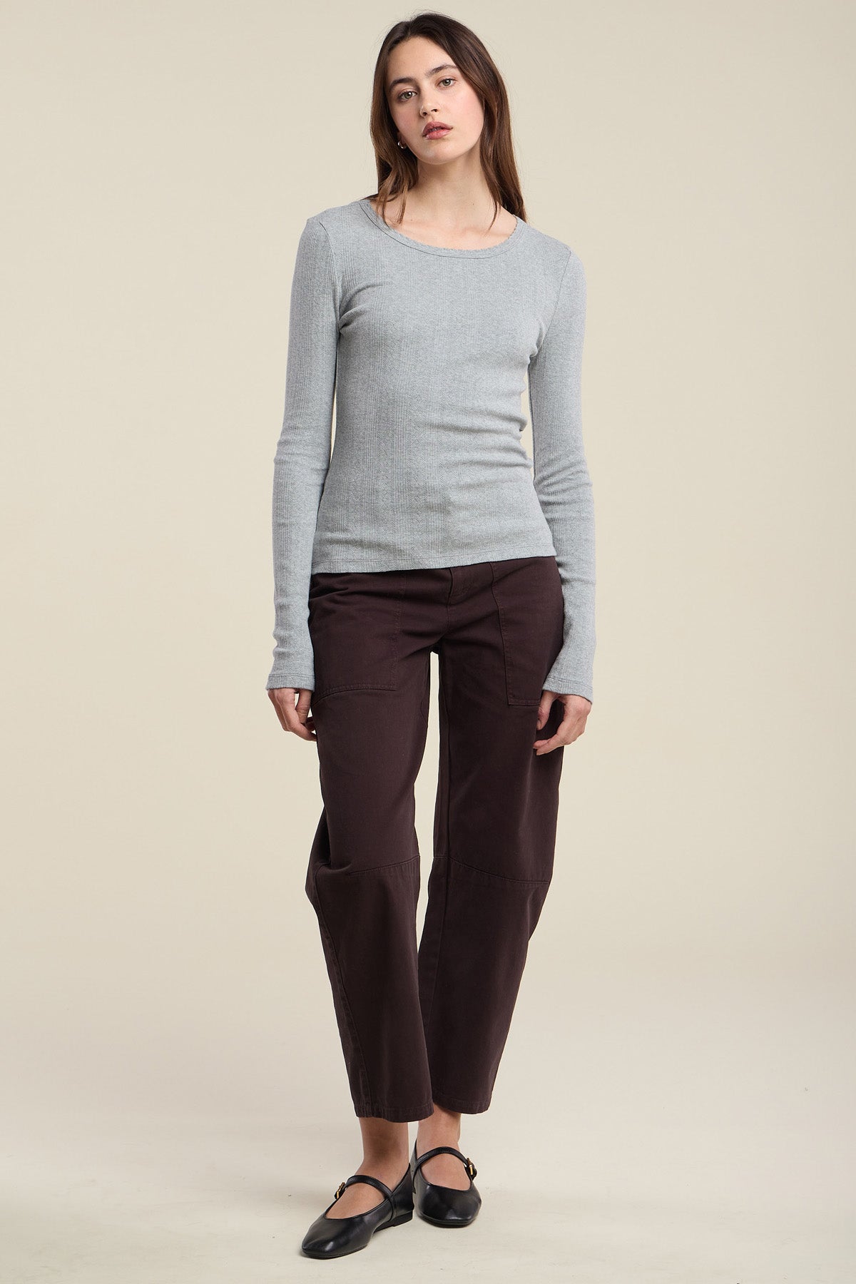 A woman stands facing forward, wearing the MIRIAM TEE by Velvet by Graham & Spencer—a long-sleeve gray top—paired with dark brown pants and black shoes, against a plain beige background.-41826782249153
