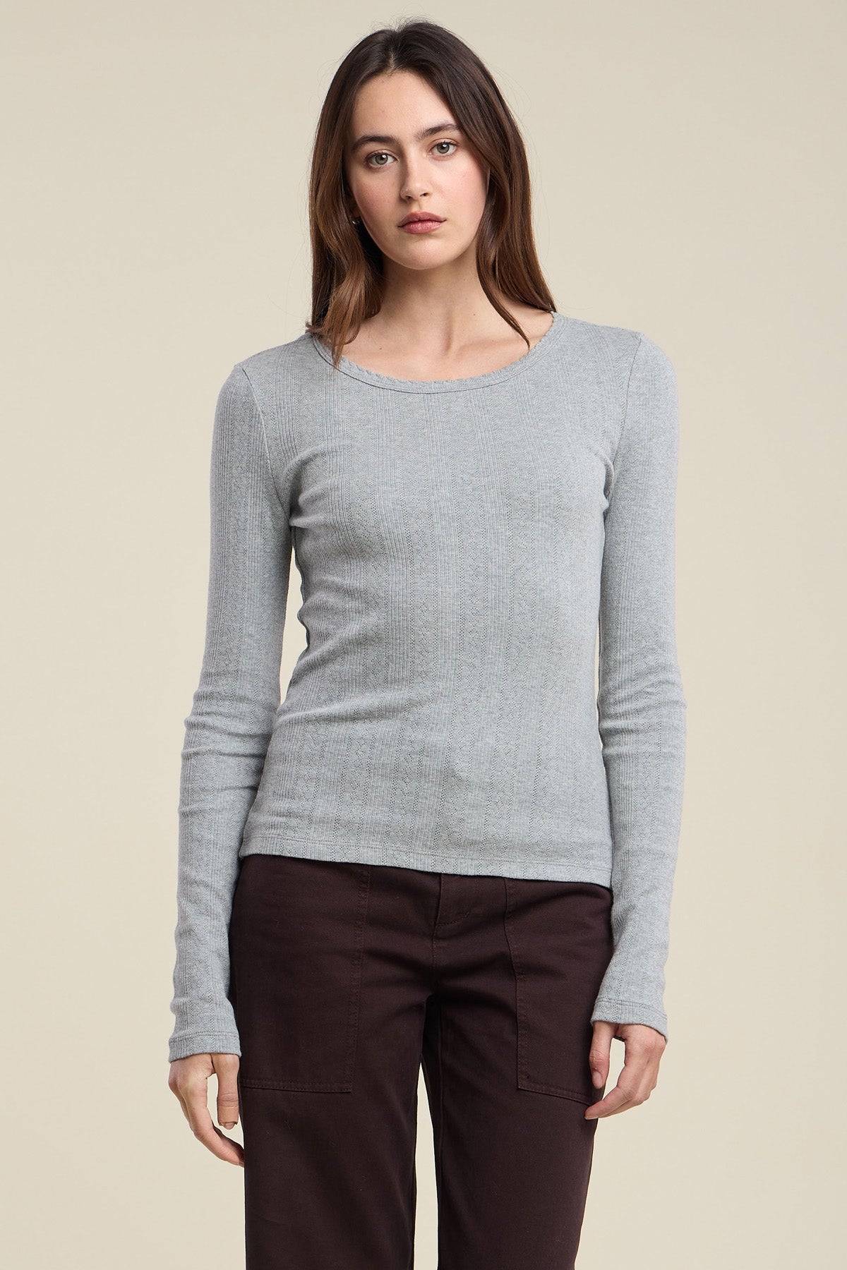 A woman stands against a plain background, facing forward with a neutral expression, wearing the light gray MIRIAM TEE by Velvet by Graham & Spencer and dark brown pants.