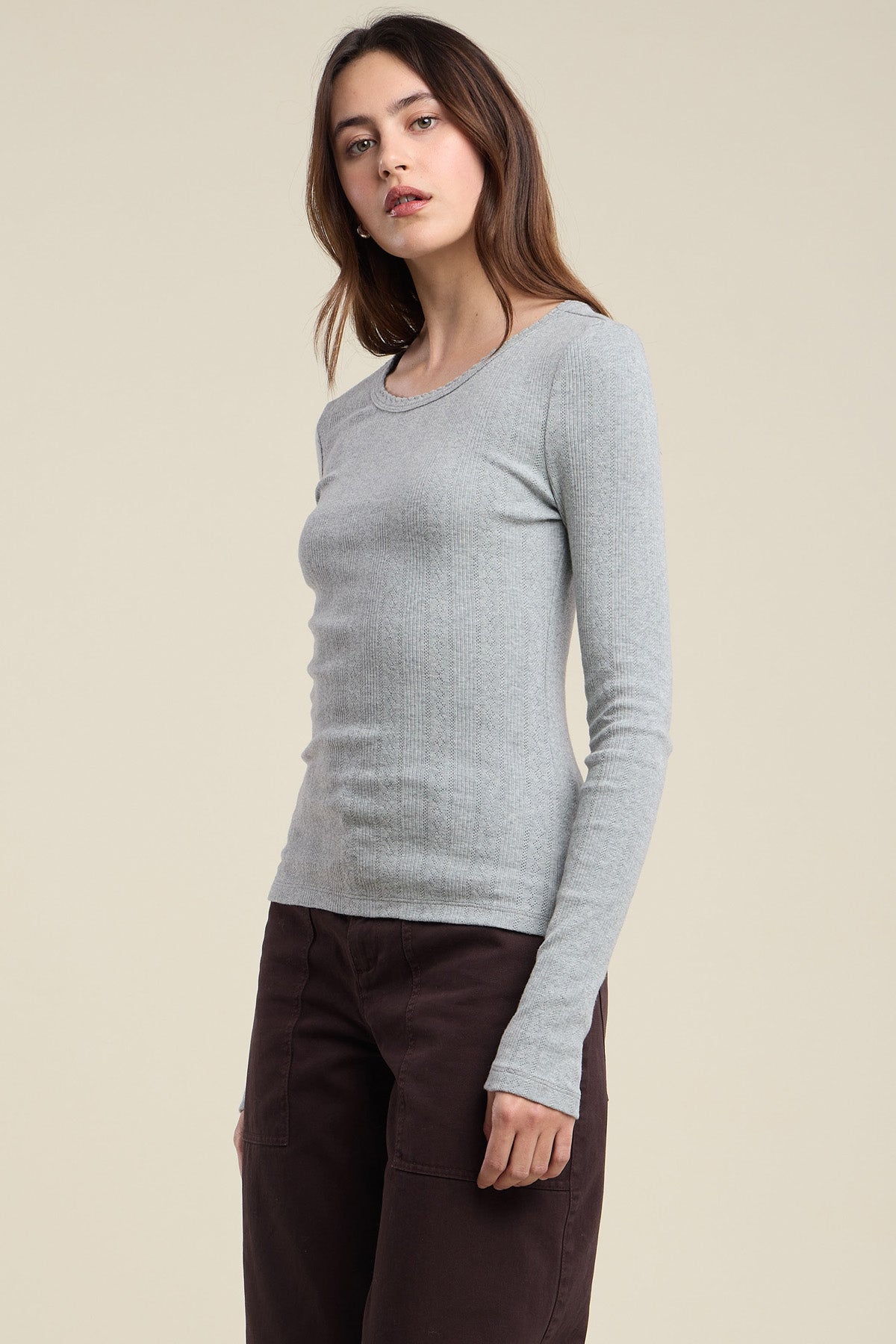 A woman wearing the MIRIAM TEE by Velvet by Graham & Spencer—a light gray, long-sleeve ribbed shirt—and brown pants stands against a plain beige background.