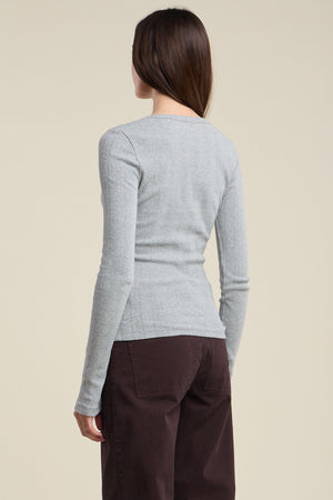 A person with long brown hair is shown from the back, wearing the MIRIAM TEE by Velvet by Graham & Spencer in light gray, paired with dark brown pants against a plain background.