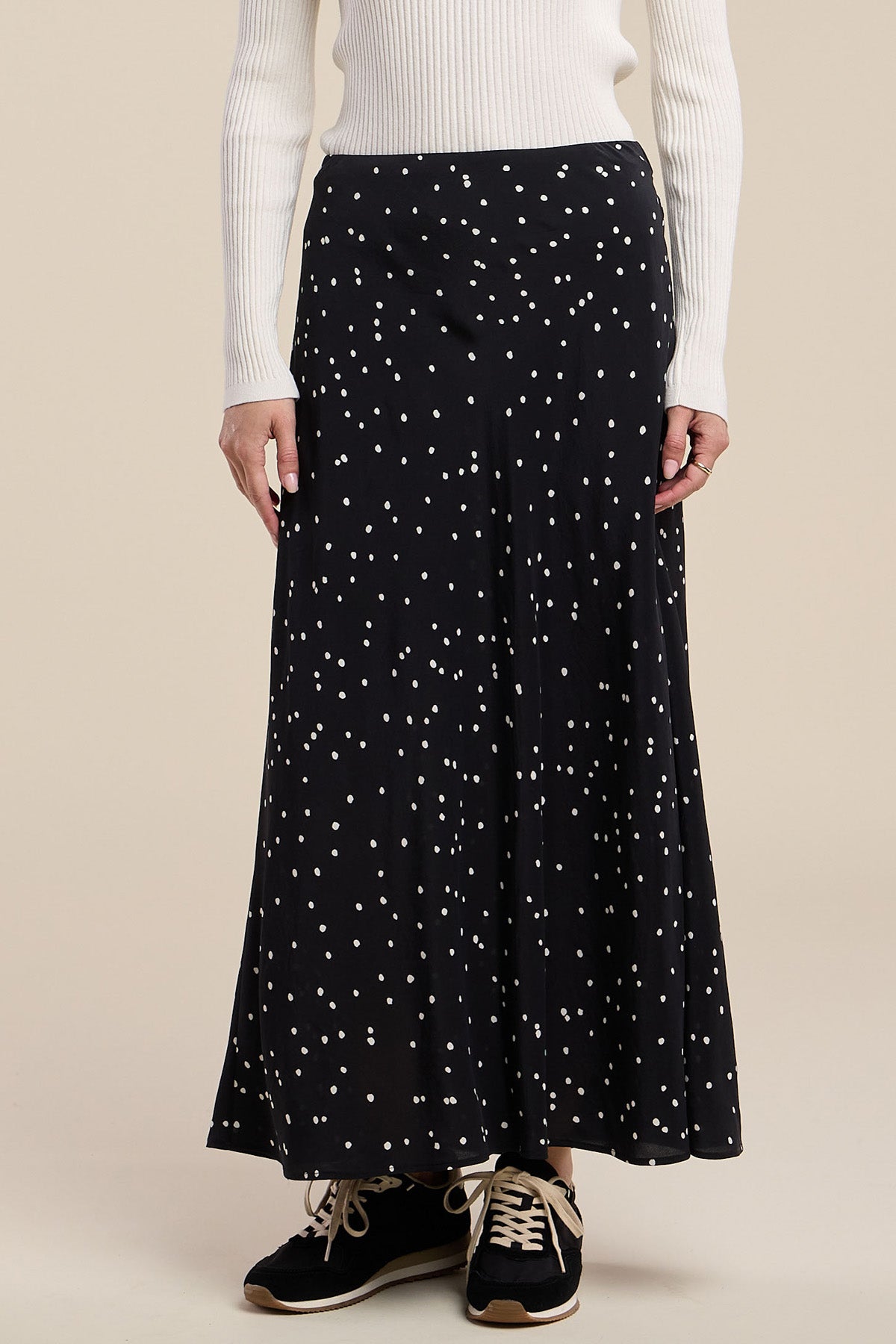 A person wearing a white ribbed sweater and the CADENCE SILK SKIRT by Velvet by Graham & Spencer—a black, ankle-length polka dot skirt in an elegant A-line silhouette—paired with black sneakers.
