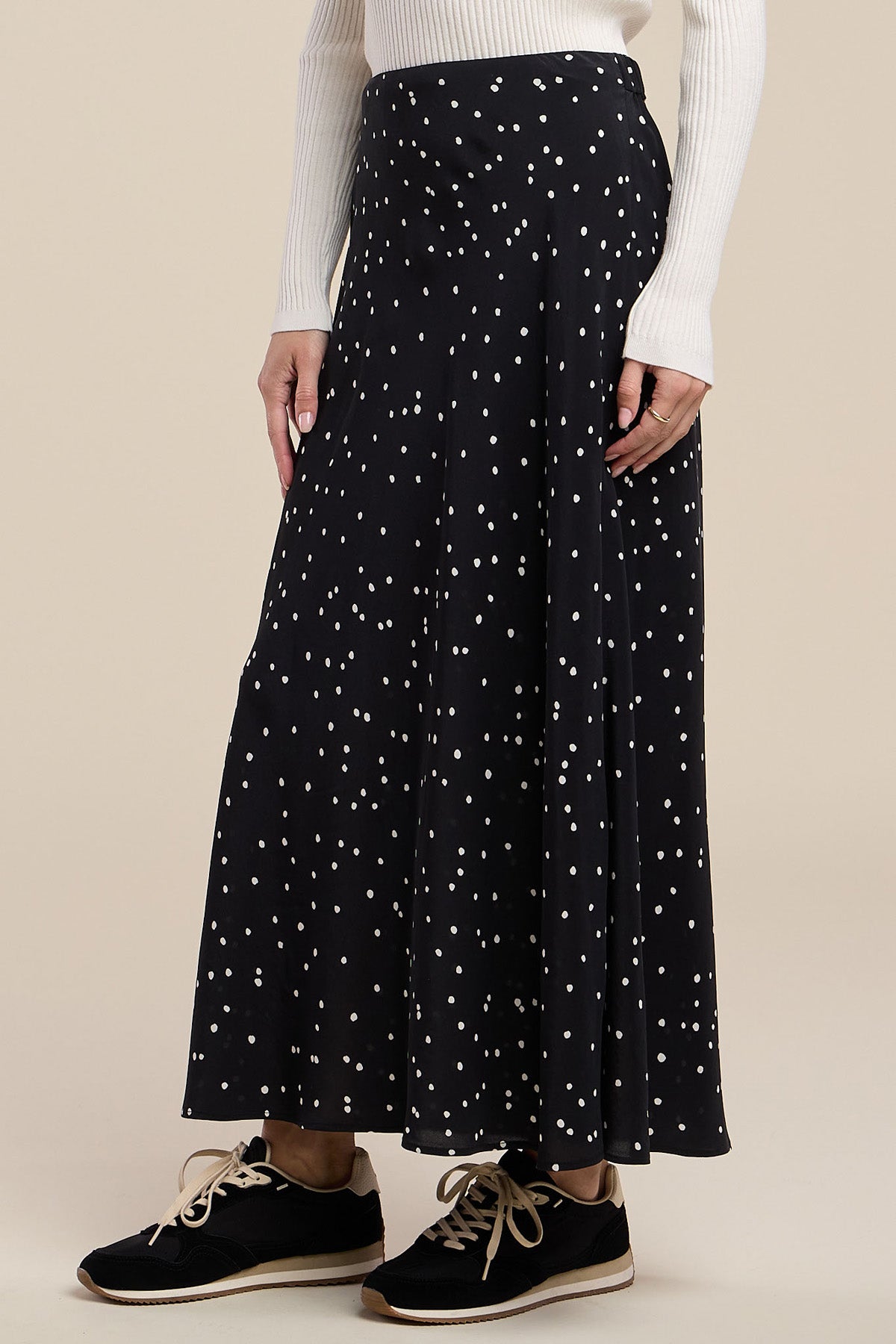 A person wearing a white ribbed long-sleeve top, the CADENCE SILK SKIRT by Velvet by Graham & Spencer—a black silk maxi skirt with white polka dots—and black sneakers with white laces, standing against a neutral background.
